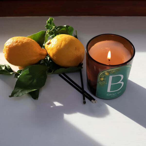 A product shot of Luminis owned Byblos candle featuring green packaging, next to two lemons and greenery.