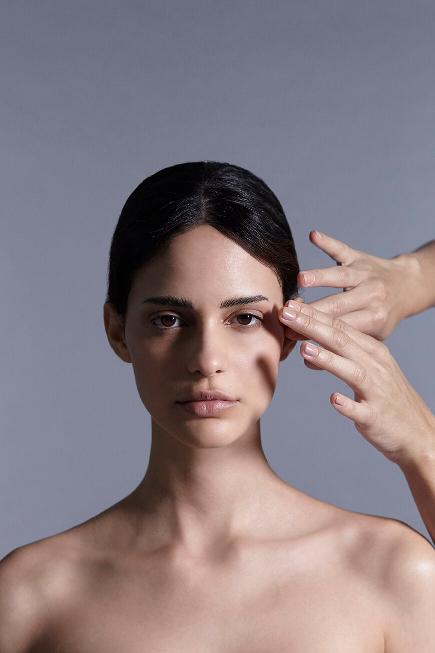 A portrait shot of a model facing directly into the camera featuring two hands from the right applying a facial massage.