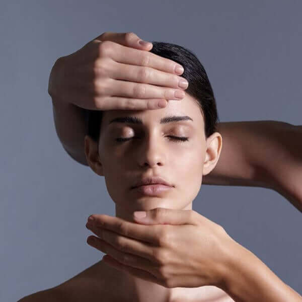 A portrait shot of a model closing her eyes featuring two hands from the right applying a facial massage.
