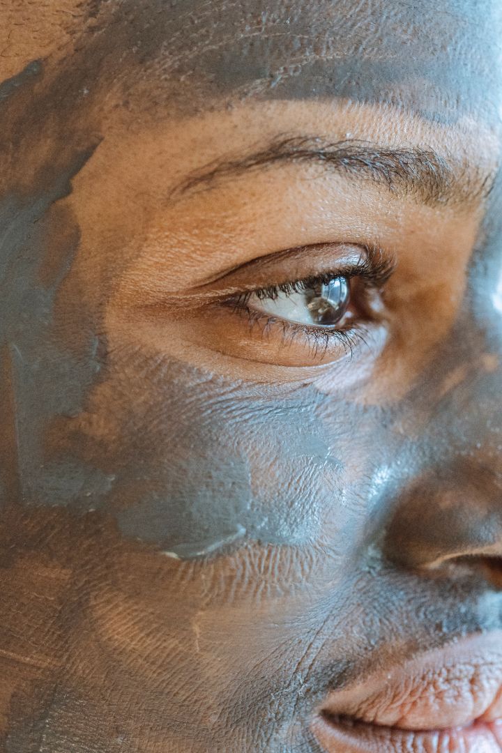 An extreme close up of a woman's face looking into the distance with a grey clay face mask on.