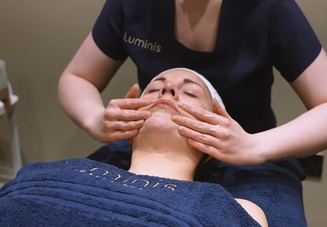 A therapist performing a facial massage on a client with Luminis branded towelling.