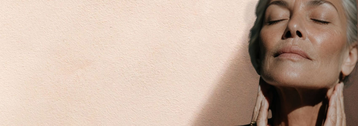 A close-up lifestyle shot of a woman with her eyes closed, gently holding her neck, set against a textured beige wall with a strong diagonal shadow.