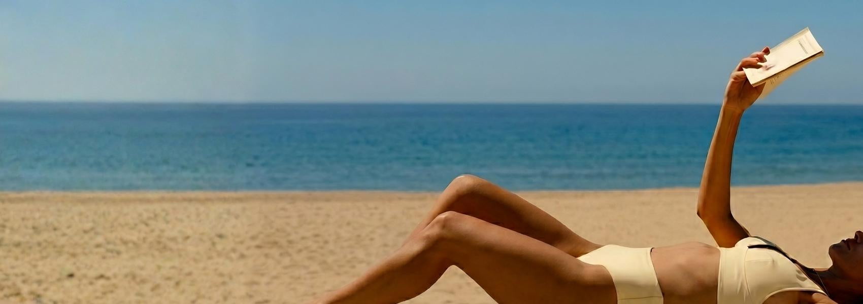 Person reading a book on a beach with clear blue sky and ocean.