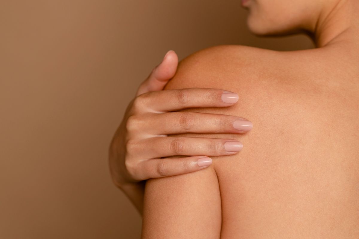 A landscape of a woman's smooth back, grabbing onto her own shoulder showcasing her manicured nails. 