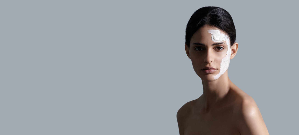 A woman with half her face covered in a white mask against a plain grey background
