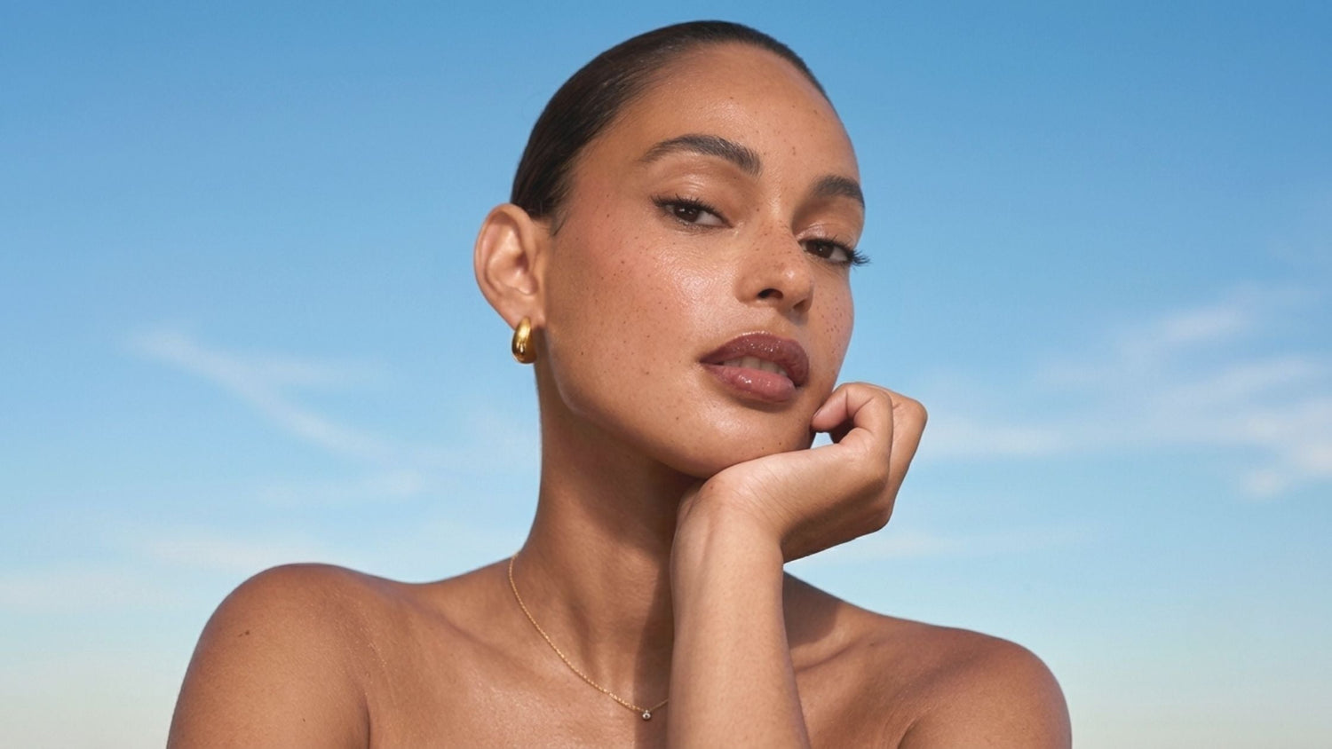 A woman with her hair slicked back, wearing gold jewellery and long, luscious lashes against a bright blue sky background 