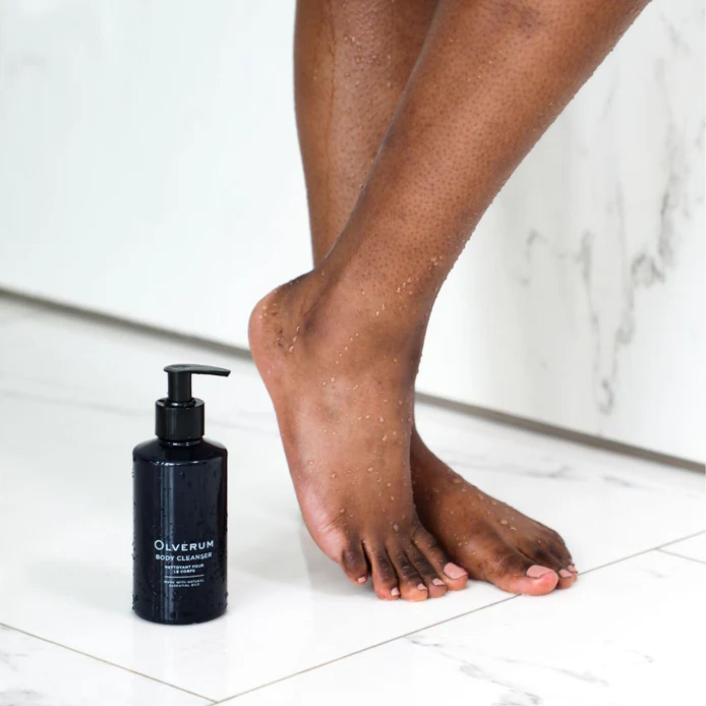 Person applying a black bottle of Oliverum foot cleanser to their feet on a white floor.