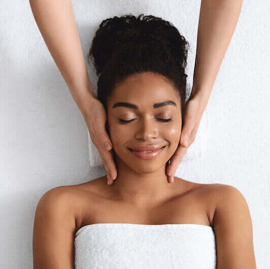A birds eye view of a lady lying down on a towel smiling whilst a therapist has both hands either side of her face.