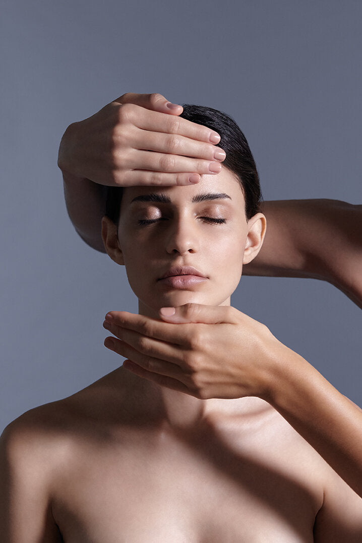 A portrait shot of a model closing her eyes featuring two hands from the right applying a facial massage. 