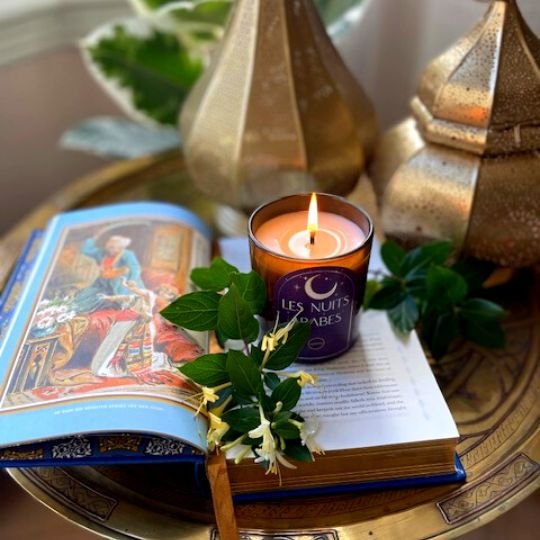 A product shot of the Les Nuits Arabes Candle whilst lit, resting on a book with gold lanterns in the background. 
