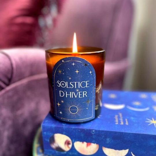 A product shot of the Solstice candle lit, with a purple velvet couch in the background. 